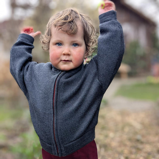 ein 2jähriges Mädchen streckt die arme nach oben. Sie trägt eine gestrickte Bomberjacke für Kinder mit Reißverschluß in grau.
