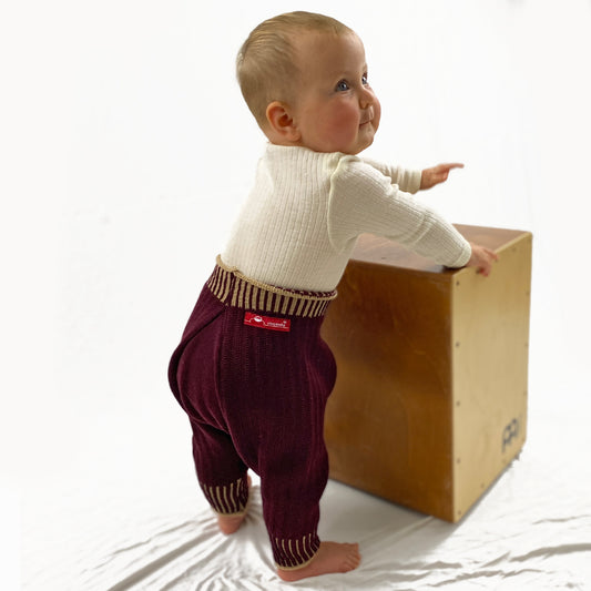 ein Baby, daß gerade mal stehen kann, trägt eine Sarouelhose auch Harems- oder Pluderhose genannt. Mit tiefgeschnittenem Schritt und extrem viel Platz für Windeln. Die Bordeaux-farbene Wollhose ist aus feinster Bio-Merinowolle gestrickt. Sie hat enganliegende hochelastischen Bündchen, die Beige Längsstreifen haben.