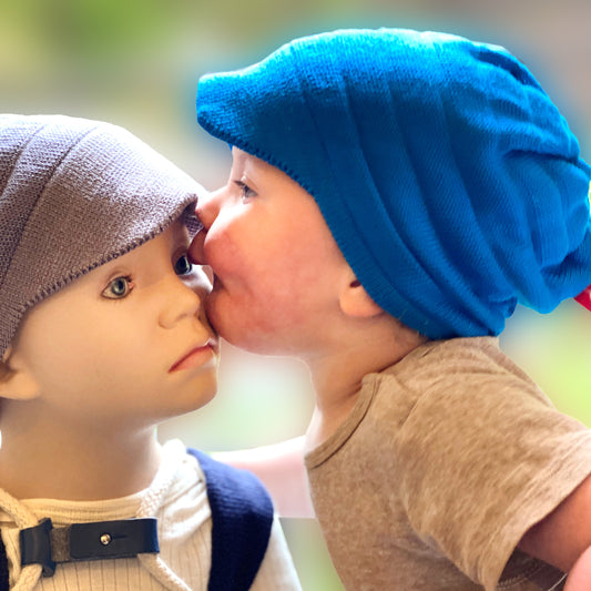 ein ca. einjähriges Kind bläst seine Backen dich auf und küßt eine Puppe es trägt eine Sommermütze mit Schirm von vincente aus Tencel/Lyocell gestrickt in einem läuchtenden Blauton.