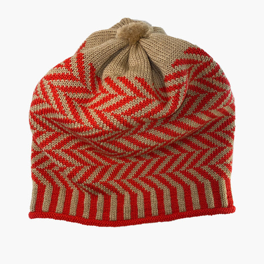 Orangerote/Karamelfarbene Beanie-Mütze aus Merinowolle mit längs gestreiften elastischem Bündchen. Die Mütze ist im klassichem Fischgrätmuster gestrickt und hat eine kleine unauffällige Bommel.