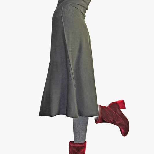 Man sieht ein Model vom Bauchnabel abwärts in einem ausgestellten über-knielangen Strickrock in Grau. Sie trägt sammetrote Schuhe mit hohen Absätzen.