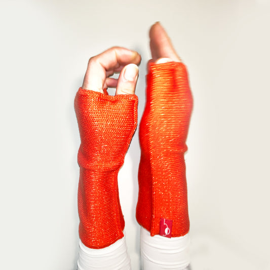 Zwei Hände präsentieren senkrecht nach oben zeigend orangerote Armstulpen mit abgenähtem Daumenloch.Der Wolle ist ein Lurexfaden beigemischt. So daß die Stulpen weihnachtlich glitzern.