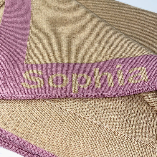 Man sieht eine zusammengefaltete Karamelfarbenen Babydecke. Der Rand ist Rosenholzfarben und mit dem Namen Sophia bestickt.