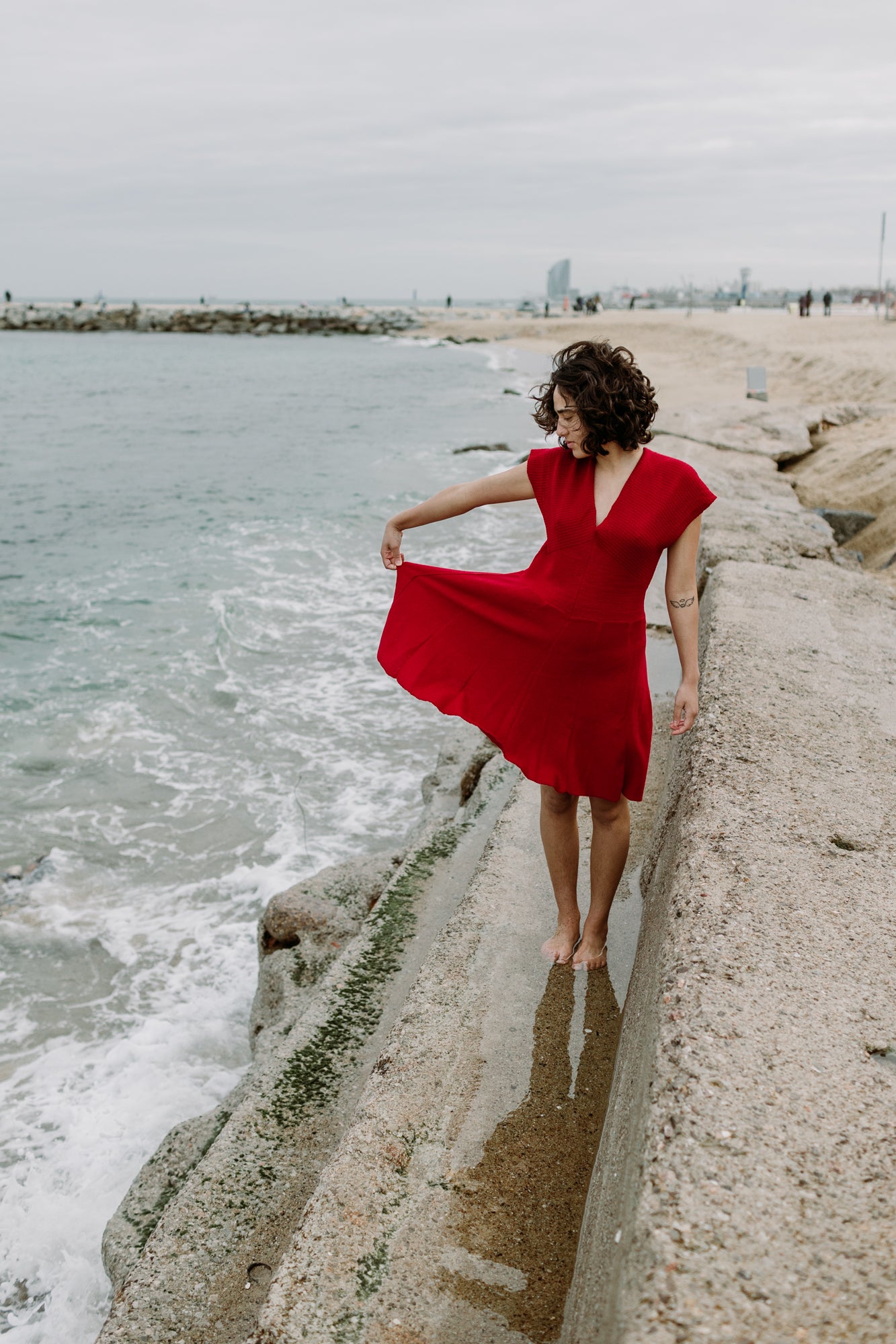 Eine junge Frau mit schwarzen Locken steht am Strand mit den Füßen im Wasser. Sie hät mit einer Hand den Rock des Kleides zur Seite, so daß man sie wie weit dieser schwingt. Es ist ein Rotes schwingendes Kleid mit angeschnittenen Ärmeln und V-Ausschnitt. aus feiner Bio-Merinowolle gestrickt von vincente