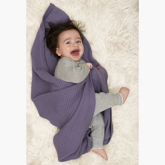 Ein kleines Baby lacht mit weit aufgesissem Mund und Augen. Es hat noch keine Zähne aber extrem viele schwarze Haare. Das baby ist in eine Lilane im Fangpatentmuster gestrickte Babydecke von vincente gehüllt