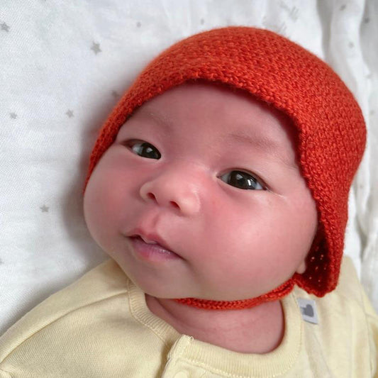 Pausbäckiges Baby guckt mit großen braunen Augen in die Kamera. Es trägt eine orange gestrickte Haube von vincente. Mit verstellbarem Bändchen zum zuknöpfen und kleinem beigen Knopf.