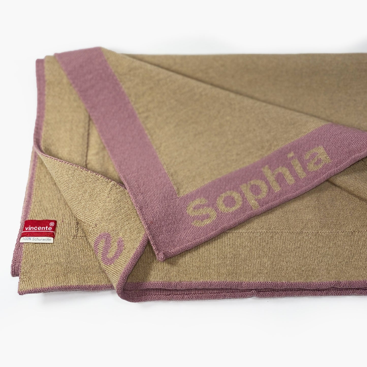 Man sieht eine zusammengefaltete Karamelfarbenen Babydecke. Der Rand ist Rosenholzfarben und mit dem Namen Sophia bestrickt.