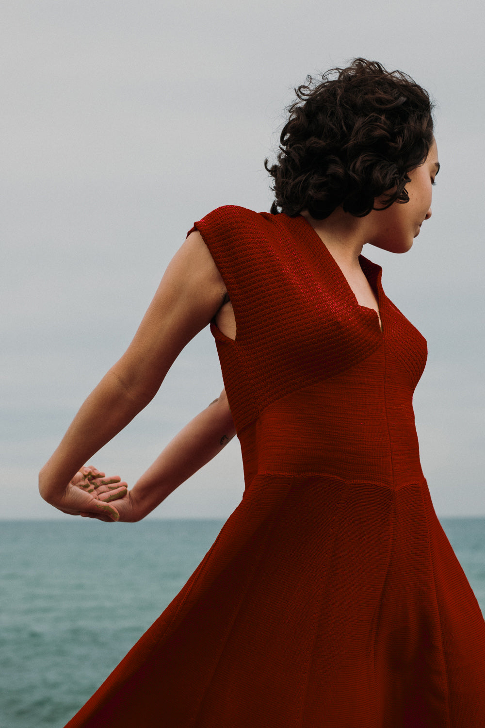 Eine junge Frau mit schwarzen Locken steht am Strand Sie trägt ein Bordeauxrotes schwingendes Kleid mit angeschnittenen Ärmeln und V-Ausschnitt. aus feiner Bio-Merinowolle gestrickt von vincente