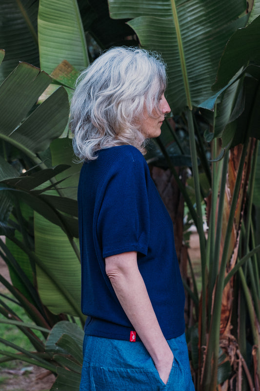 Eine Frau mit Silbergrauen Locken steht unter einer Bananenstaude. Sie trägt einen  gestrickten T-Shirt Pullover von vincente. Der Pullover hat kurze Arme und ist Royalblau
