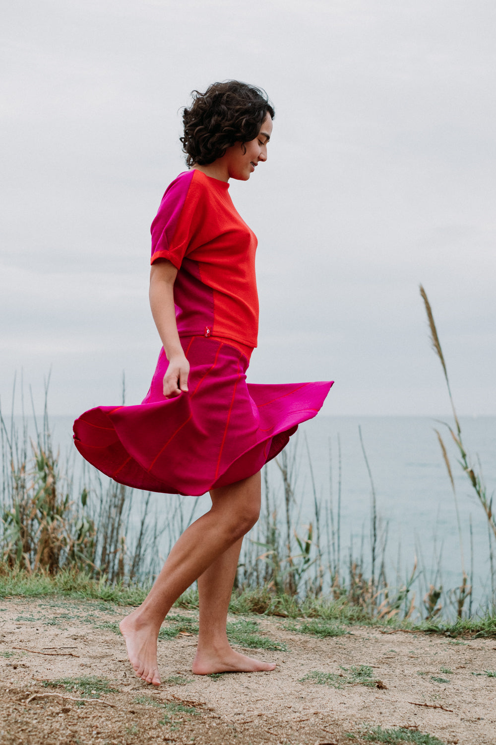 Ein junges Mädchen mit schwarzen Locken schwingt Ihren Rock am Strand. Der Rock ist Pink und hat Orange Längsstreifen. Er ist aus Merinowolle gesrtrickt. Auch das Oberteil von vincente ist in den 2 Farben gestrickt.