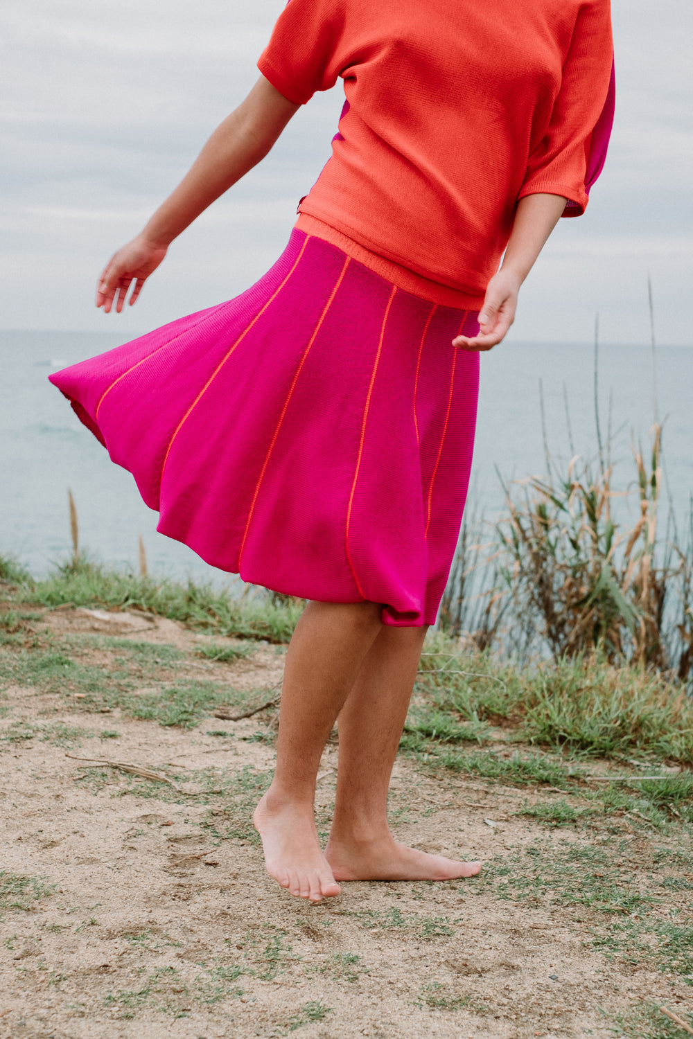 Ein junges Mädchen mit schwarzen Locken schwingt Ihren Rock am Strand. Der Rock ist Pink und hat Orange Längsstreifen. Er ist aus Merinowolle gesrtrickt. Auch das Oberteil von vincente ist in den 2 Farben gestrickt.