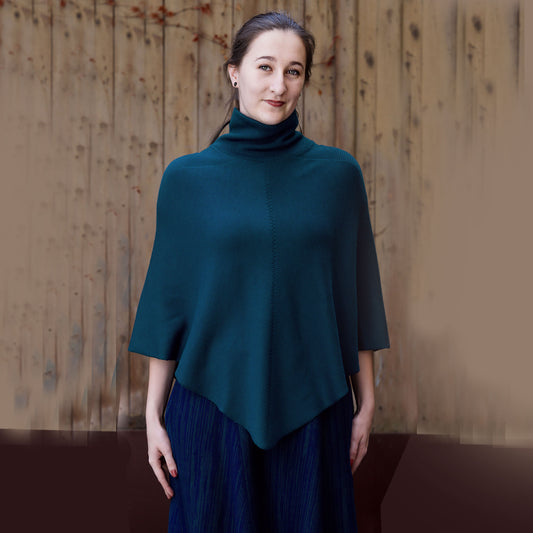 Eine Junge Frau trägt ein Petrolfarbenes, gestricktes Cape mit Rollkragen