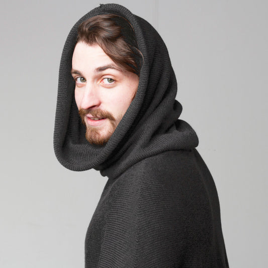 Ein junger Mann mit Don Quichotte Bart hat einen den großen U-bootkragen des grauen gestrickten Capes über den Kopf gestülpt.