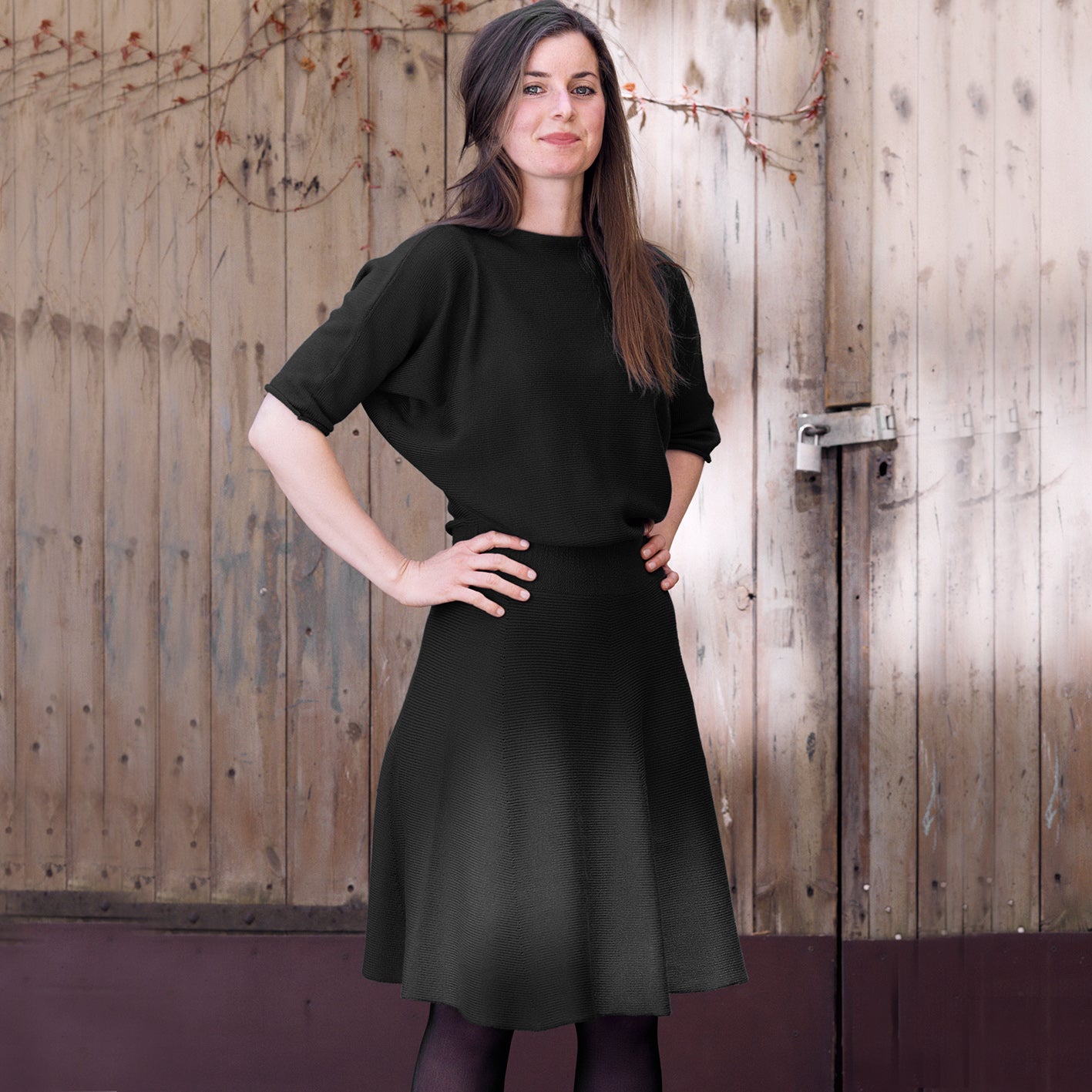 Eine Junge Frau mit langen braunenHaaren trägt ein schwarzes, tailliertes Strickkleid mit 3/4langen Fledermausärmeln und ausgestellten Rock. Sie steht vor einem hölzernen Scheunentor.