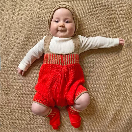 gut gelauntes Babygestrickte streckt alle viere von sich und trägt eine kurze Hose aus feinster Bio-Merinowolle mit tiefen Schritt und viel Platz für Windeln. Die Hose von vincente hat hochelastische Hosenträger und ist Orange. Die Hosenträger sind beige so wie die längsstreifen in dem geraffsten Bund und Beinabschlüssen. Auf dem Kopf trägt es eine gestrickte beige Haube und orangerote Söckchen an den Füßen.