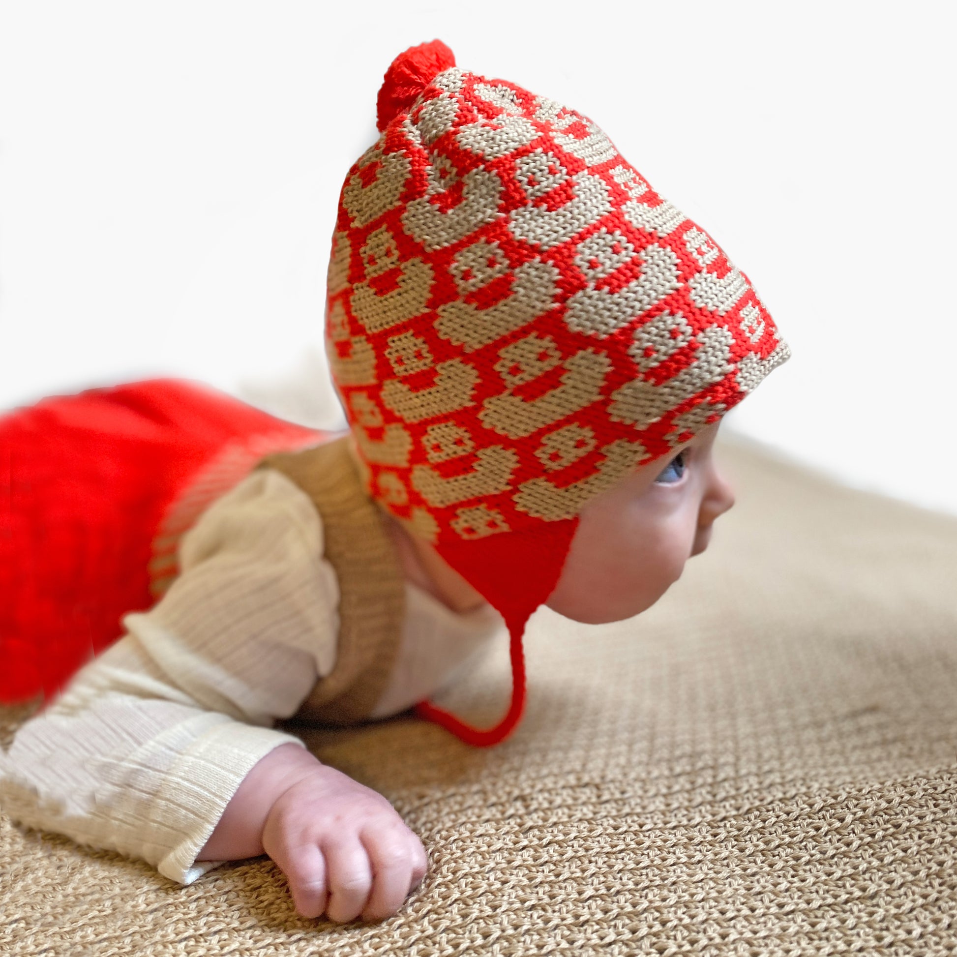 Ein kleines Baby, schafft es seinen Kopf zu heben und guckt neugierig in die Welt. Es trägt eine Orangerot/Beige Mütze mit einem fortlaufenden Muster mit kleinen Enten. Die Babymütze hat eine Bommel und angestrickte Ohrläppchen mit Bändern zum festbinden. Sie ist aufwendig der Kopfform nachgestrickt und paßt sich sehr gut an. Das rote Label mit der kleinen Ente ist hinten angenäht.