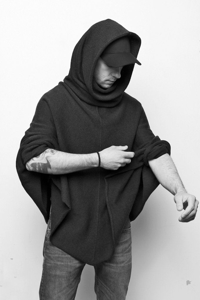Ein junger Mann mit Don Quichotte Bart hat einen den großen U-bootkragen des schwarzen gestrickten Capes über den Kopf gestülpt. Er schopped die Ärmel hoch und zeigt seine tätowierten Arme.