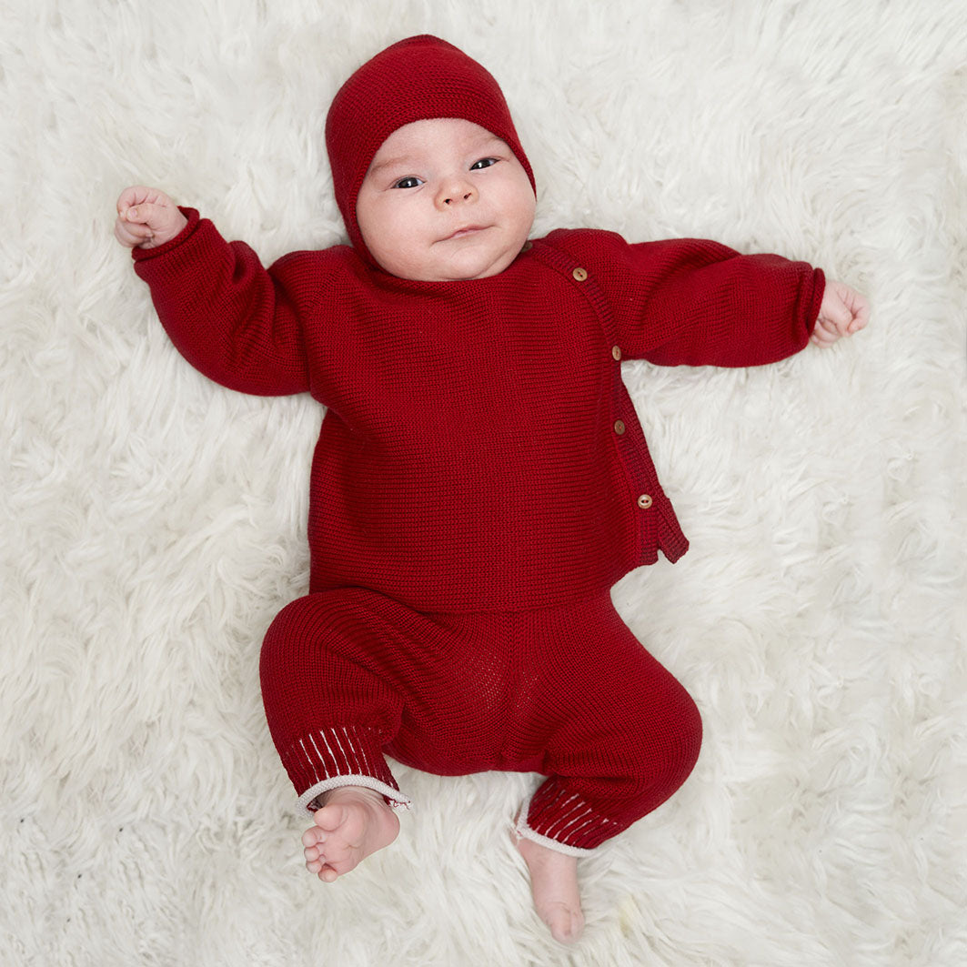 Kleines Baby ist von oben bis unten in roter gestrickter vincente-Kleidung angezogen. Es trägt eine Haube eine seitlich geknöpfte Strickjacke und eine Babyhose.