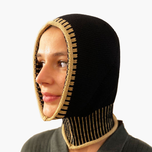 Eine Junge Frau guckt aus einer Schwarzen, gestrickten Mütze wie eine Sturmhaube. Am Hals und um das Gesicht mit elastischem längsgestreiften Bund in Karamel.