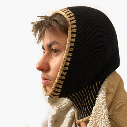 Eine junger Mann guckt aus einer Schwarzen, gestrickten Mütze wie eine Sturmhaube. Am Hals und um das Gesicht mit elastischem längsgestreiften Bund in Karamel.