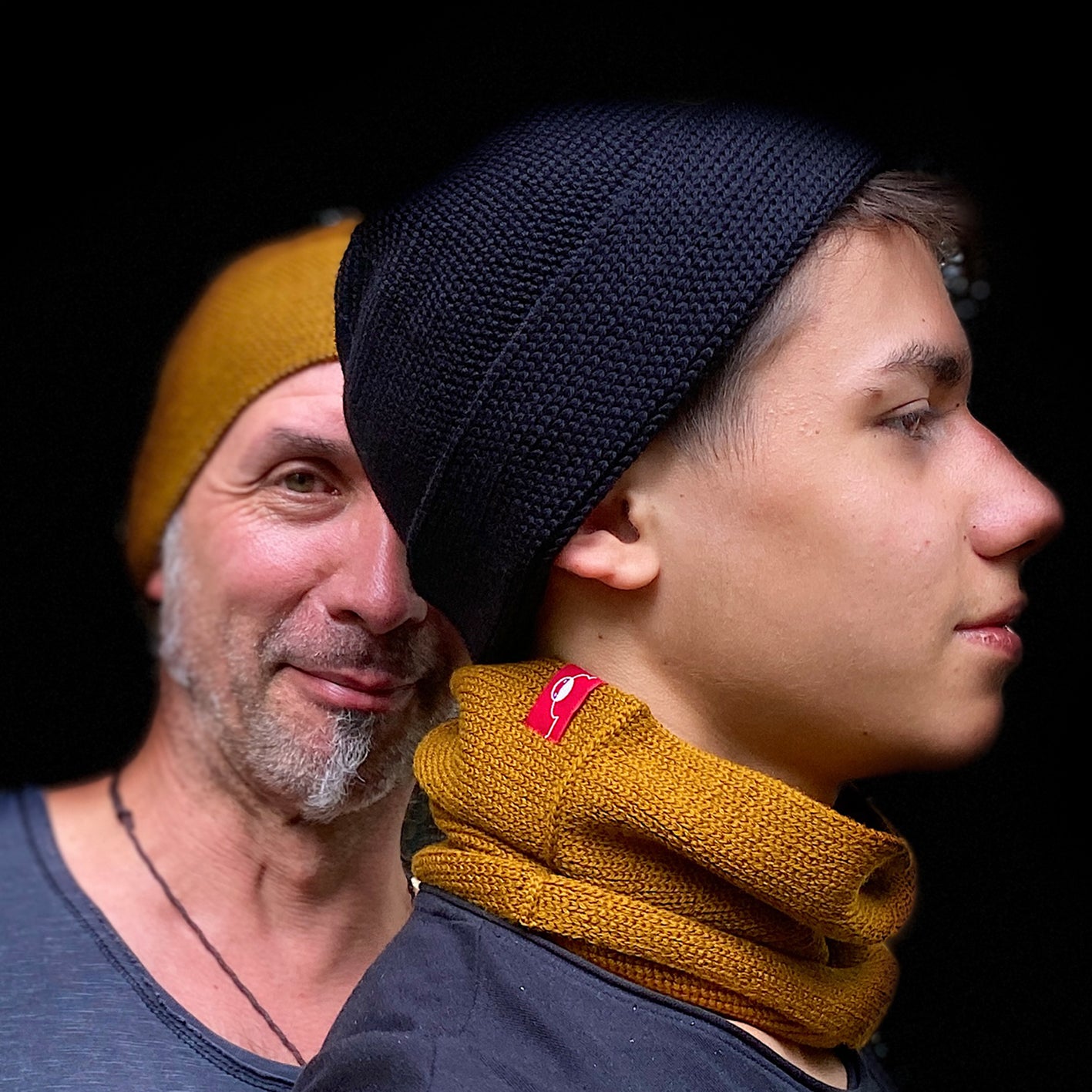 Vater und Sohn tragen beide Beanie-Mützen. Der Vater mit grauem 3Tage Bart in Bronce, der Sohn in Schwarz. Die Wollmützen sind aus Merinowolle quer in Kopf-Form gestrickt. Das Fangpatentmuster bedeutet, daß Innen ein Knötchenmuster und außen ein klasiches Rechte Maschenmuster zu sehen ist. Wenn man die Mütze umkrempelt ist jeweils das andere Muster zu sehen. Das rote vincente-Label mit der kleinen Ente ist am Rand angenäht. Die Mütze kann insideout getragen werden.