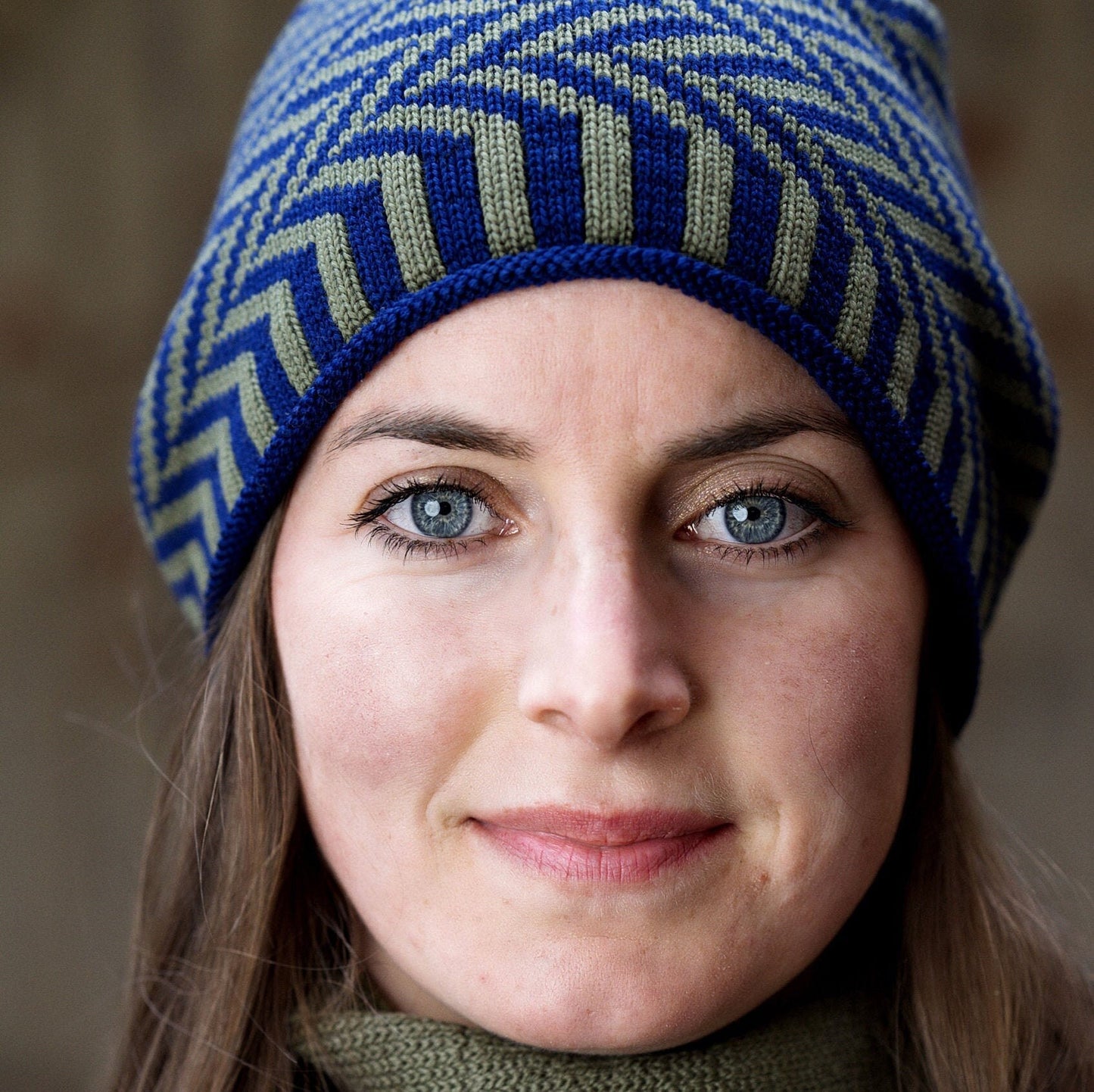 Eine junge Frau mit großen blauen Augen trägt eine Royalblaue und olivefarbene Beanie-Mütze aus Merinowolle mit längs gestreiften elastischem Bündchen. Die Mütze ist im klassichem Fischgrätmuster gestrickt und hat eine kleine unauffällige Bommel.