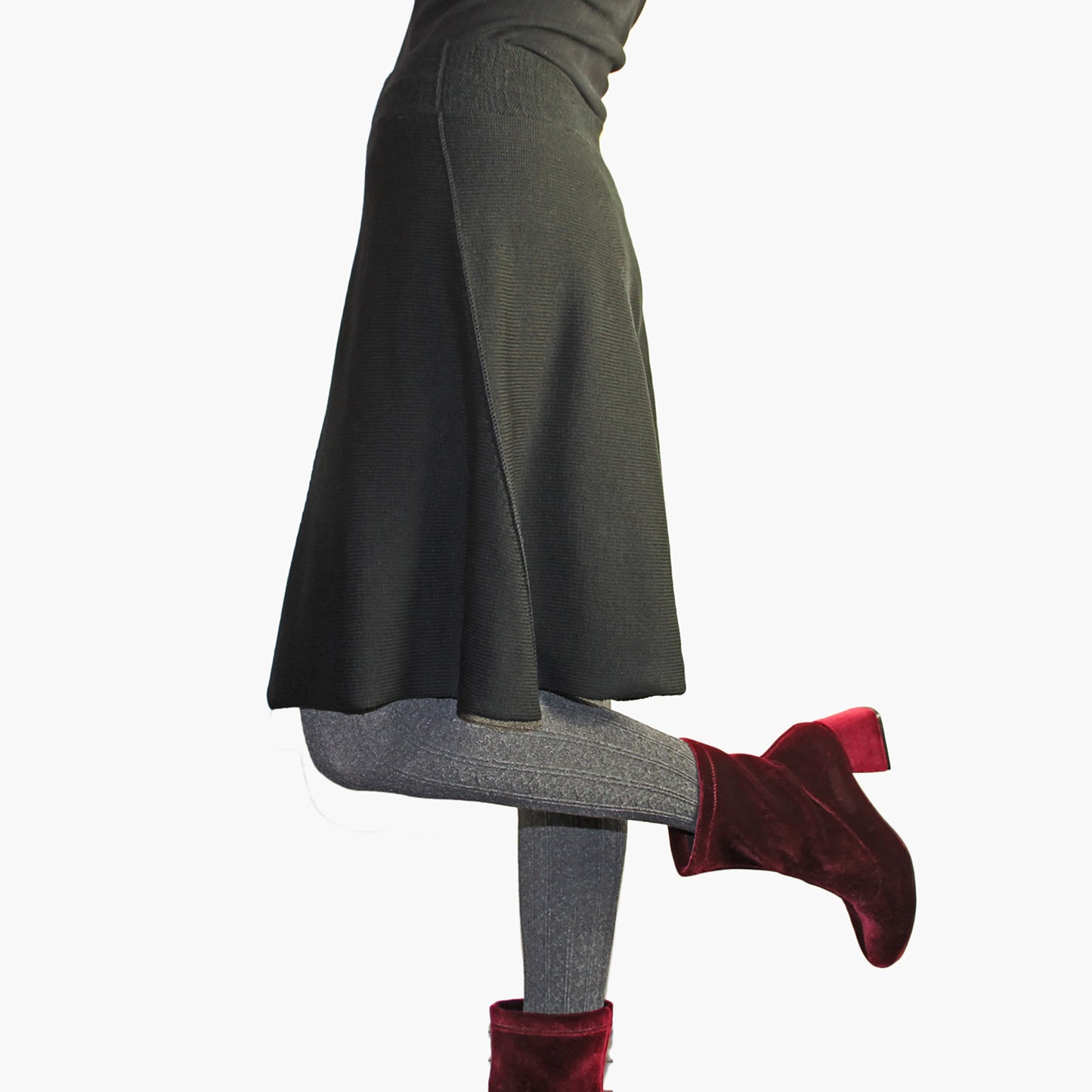 Man sieht ein Model vom Bauchnabel abwärts in einem ausgestellten knielangen Strickrock in Schwarz. Sie trägt sammetrote Schuhe mit hohen Absätzen.