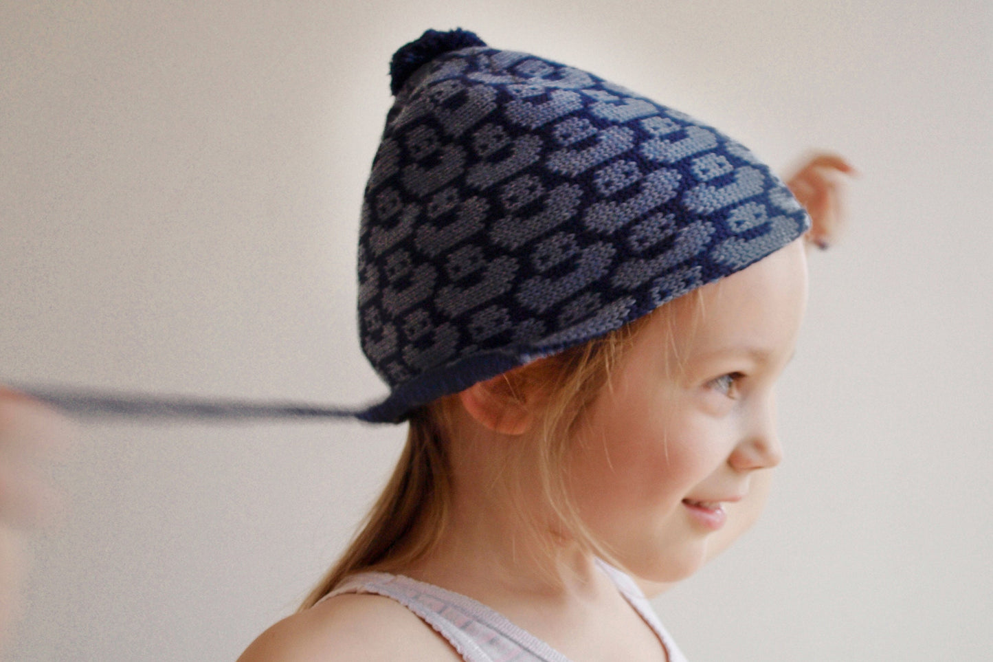 ein 6-jähriges Mädchen trägt eine Roaylblau/Jeansblaue Mütze mit einem fortlaufenden Muster mit kleinen Enten. Die Babymütze hat eine Bommel und angestrickte Ohrläppchen mit Bändern zum festbinden. Sie ist aufwendig der Kopfform nachgestrickt und paßt sich sehr gut an. Das rote Label mit der kleinen Ente ist hinten angenäht.