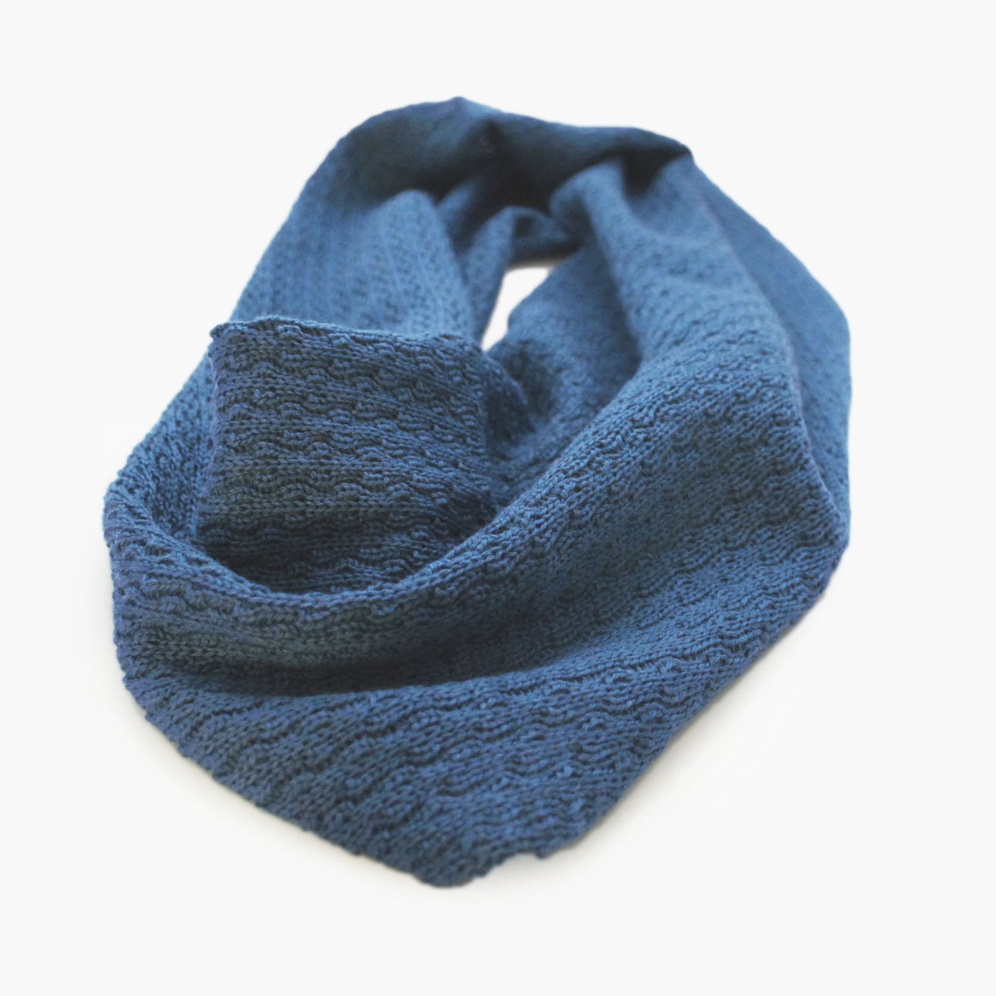 Blauer, gestrickter Loop im Fangpatentmuster. Eine Seite sieht glatt rechts mit kleinen Wellen gestrickt aus, die andere Seite sieht aus wie kleine Muscheln. Der Wollschal ist zu einem Haufen drapiert, so sieht man den weichen Fall.