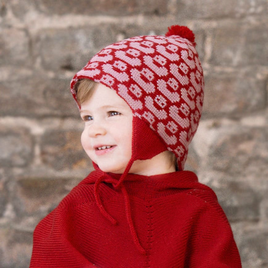 Lachendes Kind trägt Rot/Rosa Mütze mit einem fortlaufenden Muster mit kleinen Enten. Die Babymütze hat eine Bommel und angestrickte Ohrläppchen mit Bändern zum festbinden. Sie ist aufwendig der Kopfform nachgestrickt und paßt sich sehr gut an. Das rote Label mit der kleinen Ente ist hinten angenäht.