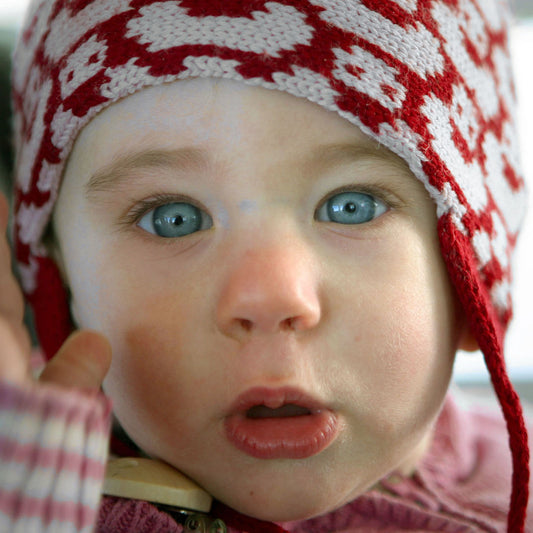 Ein Baby guckt neugierig in die Welt. es trägt eine Rot/Wollweiße Mütze mit einem fortlaufenden Muster mit kleinen Enten. Die Babymütze hat eine Bommel und angestrickte Ohrläppchen mit Bändern zum festbinden. Sie ist aufwendig der Kopfform nachgestrickt und paßt sich sehr gut an. Das rote Label mit der kleinen Ente ist hinten angenäht.
