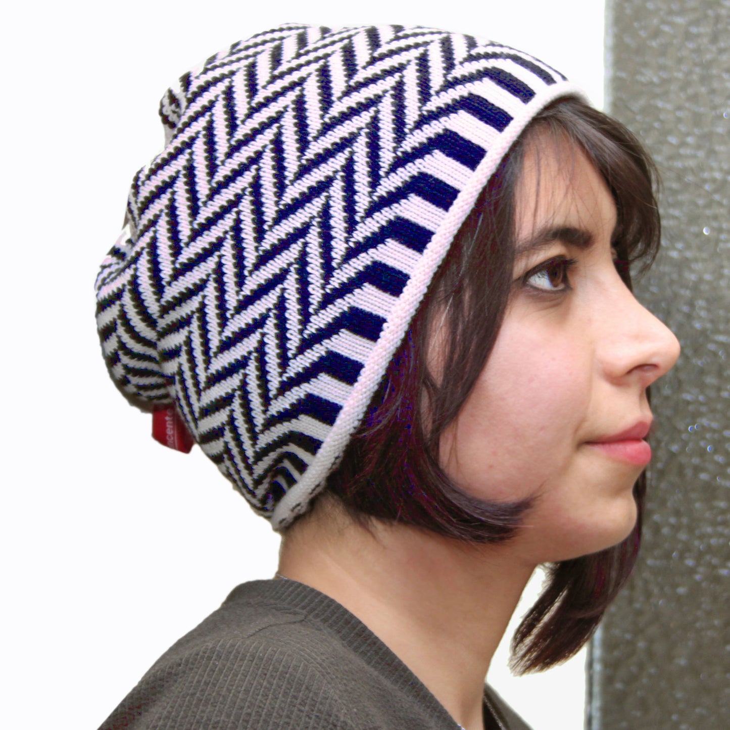 Eine junge Frau trägt eine Weiß und Royalfarbene Beanie-Mütze aus Merinowolle mit längs gestreiften elastischem Bündchen. Die Mütze ist im klassichem Fischgrätmuster gestrickt und hat eine kleine unauffällige Bommel.