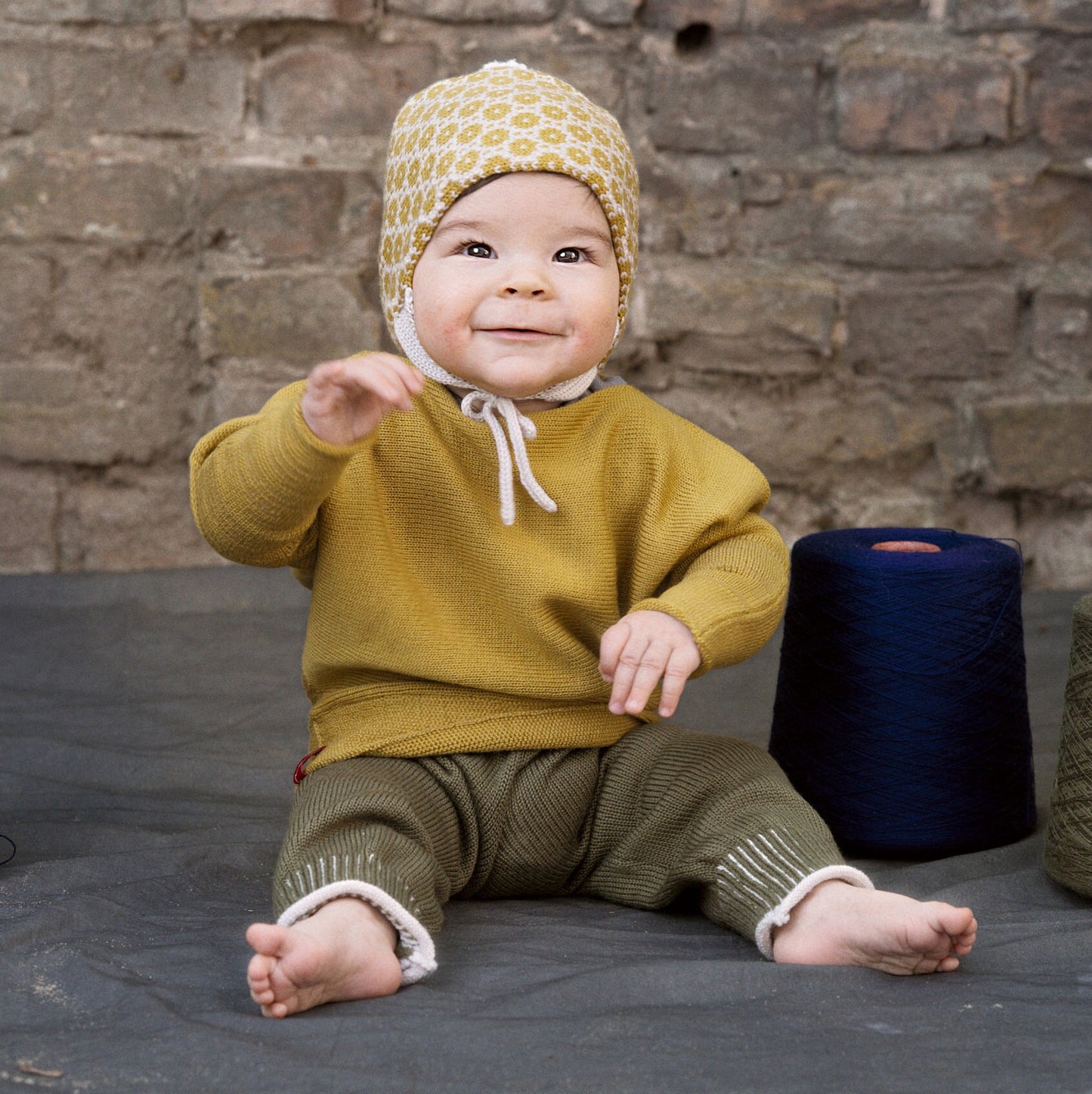 Ein Baby sitzt auf einer Decke. Neben ihm stehen Wollkonen. Es ist von oben bis unten in Strickkleidung von vincente angezogen