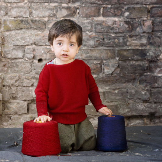 Ein kleiner Junge spielt mit Wollkonen. Er trägt einen Roten gestrickten Pullover mit angeschnittenen Ärmeln. Der Pullover hat einen elastichen Bund und lange elastische Ärmelbündchen.