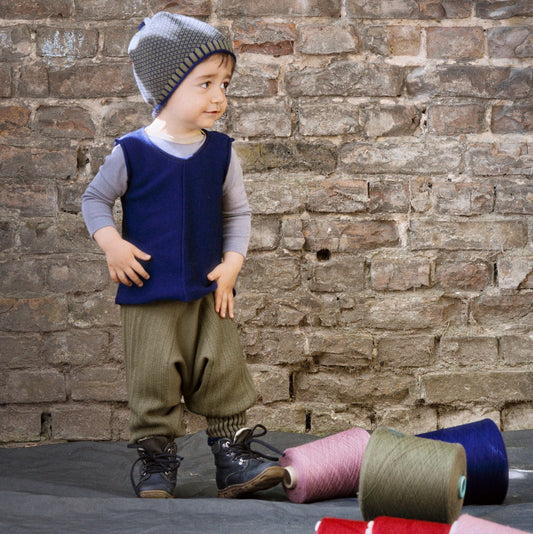 Ein ca 1,5 jähriger Junge spielt mit Wollkonen wie mit Bauklötzen. Er lächelt schelmisch, weil er seinen Turm gerade umgeworfen hat. Der Junge ist in Strickwaren von vincente gekleidet. Er trägt eine karierte Mütze einen Pullunder in royalblau und eine Sarouelhose in Olivegrün