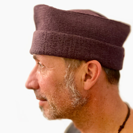 Ein Mann trägt eine Auberginefarbene, klassische, gestrickte Matrosen- oder Schiffchenmütze. Mit Rotem vincente-Label mit der weißen Ente.