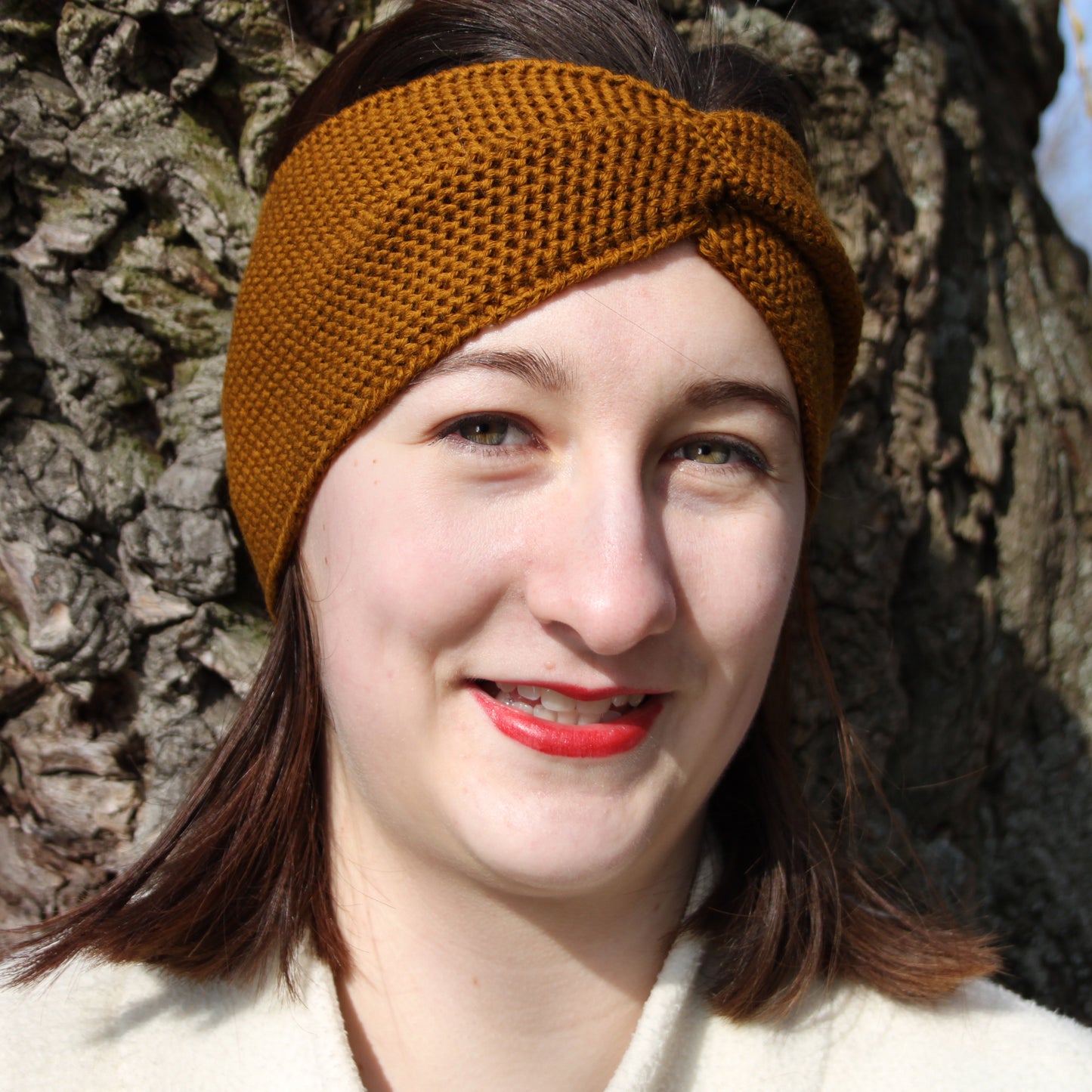 Eine Junge Frau trägt ein Bronce-farbenes Stirnband mit einer Turban-ähnlichen Verdrehung. Gestrickt im Doubleface Patentmuster, außen Knötchenstruktur und Innen sieht es wie ein klassisch rechts gestricktes Muster aus.