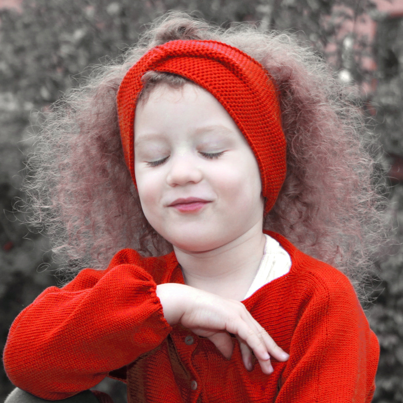 ein kleines Mädchen mit langen lockigen Haaren trägt ein Orangerotes Stirnband mit einer Turban-ähnlichen Verdrehung. Gestrickt im Doubleface Patentmuster, außen Knötchenstruktur und Innen sieht es wie ein klassisch rechts gestricktes Muster aus.