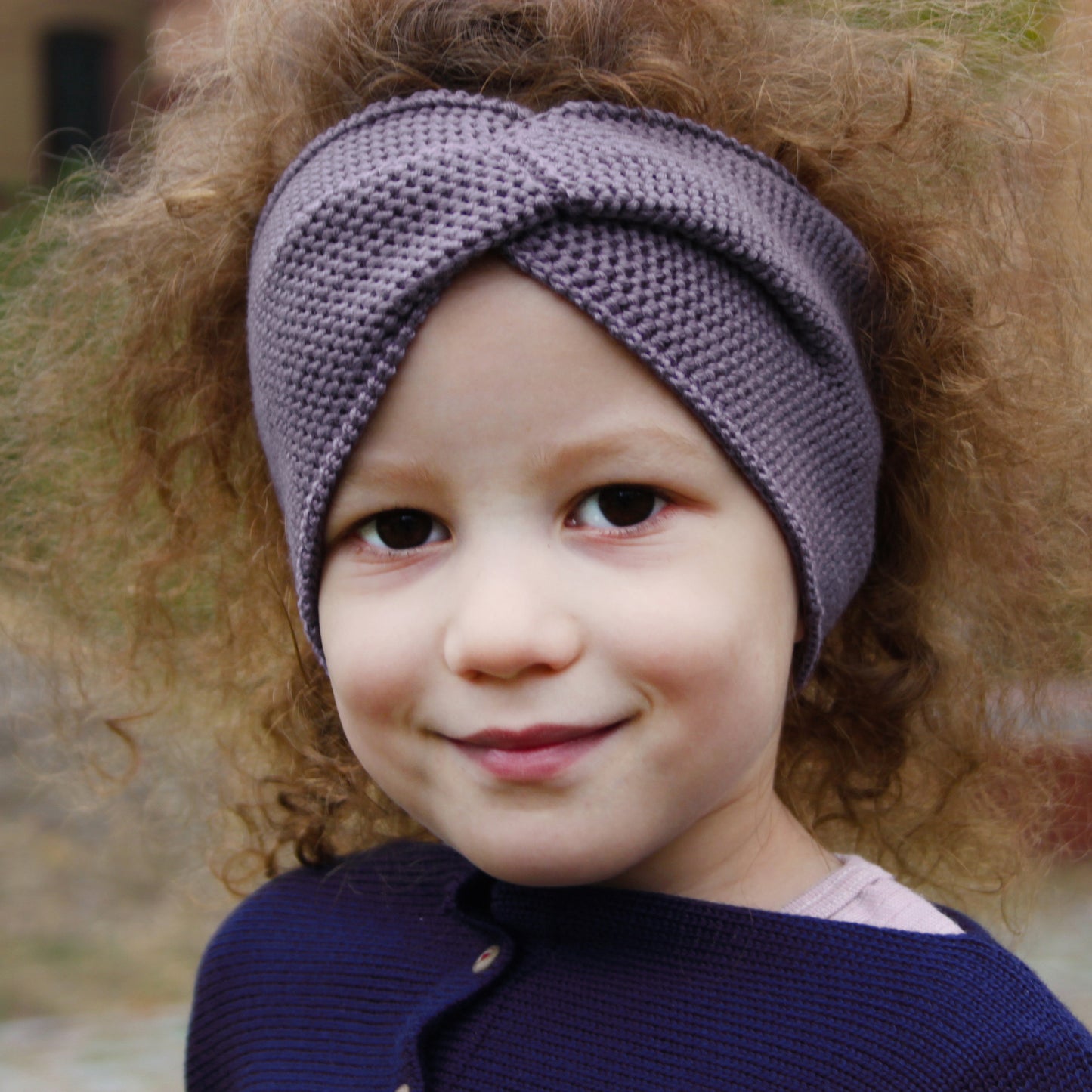 Ein kleines Mädchen mit wuscheligen Haaren trägt ein Lila Stirnband mit einer Turban-ähnlichen Verdrehung. Gestrickt im Doubleface Patentmuster, außen Knötchenstruktur und Innen sieht es wie ein klassisch rechts gestricktes Muster aus.