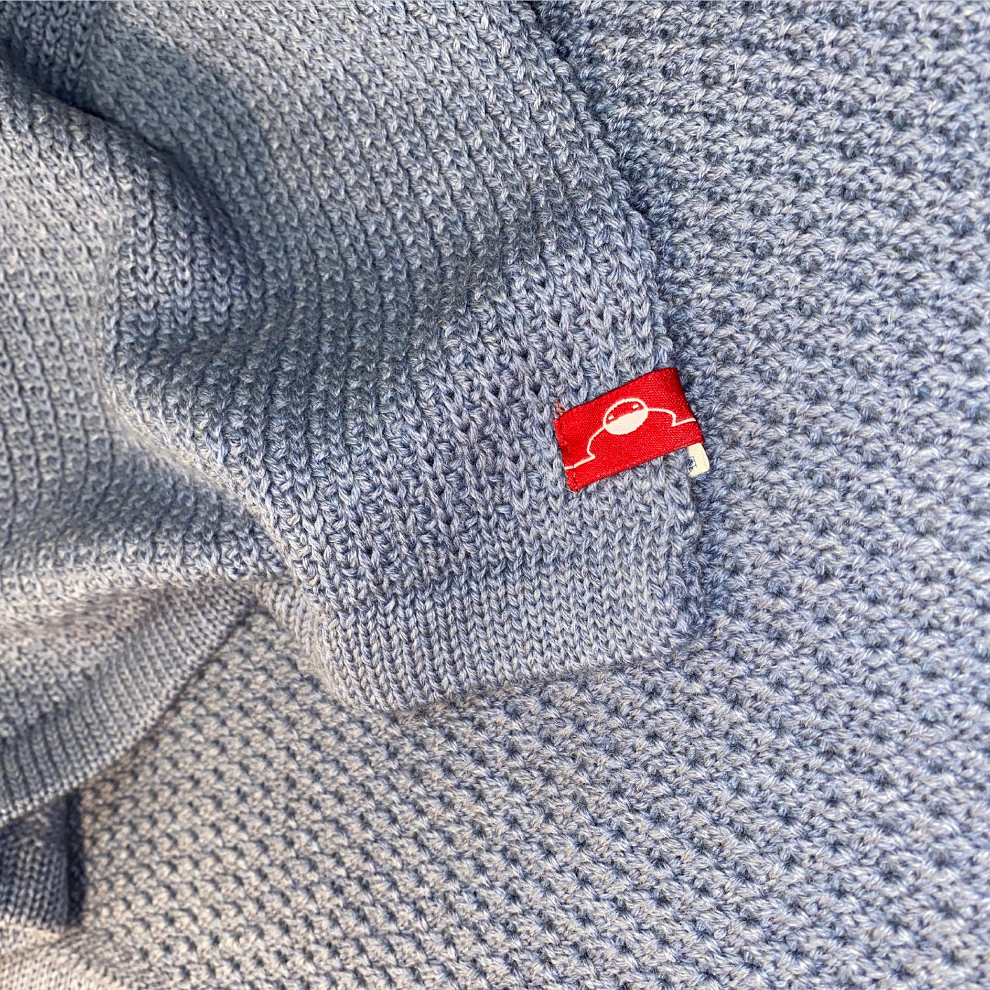 Closeup einer gestrickten Babydecke in Graublau. Man sieht das rote Logo von vincente und das das strickmuster auf der linken Seite Muschelartig ist.