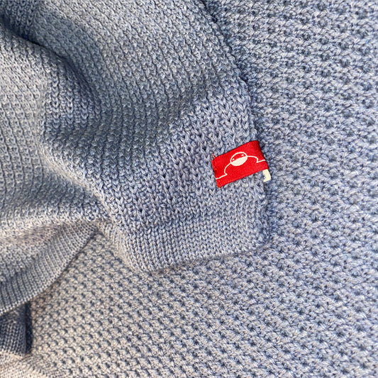 Closeup einer gestrickten Babydecke in Graublau. Man sieht das rote Logo von vincente und das das strickmuster auf der linken Seite Muschelartig ist.