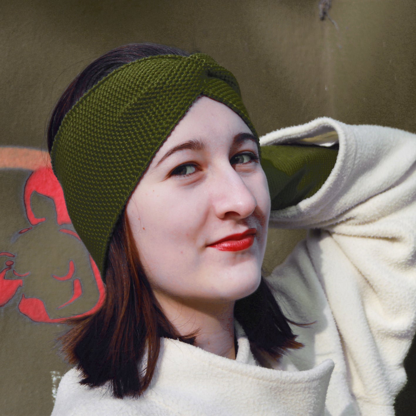 Eine Junge Frau trägt ein Olive- Farbenes Stirnband mit einer Turban-ähnlichen Verdrehung. Gestrickt im  Doubleface Patentmuster, außen Knötchenstruktur und Innen sieht es wie ein klassisch rechts gestricktes Muster aus.