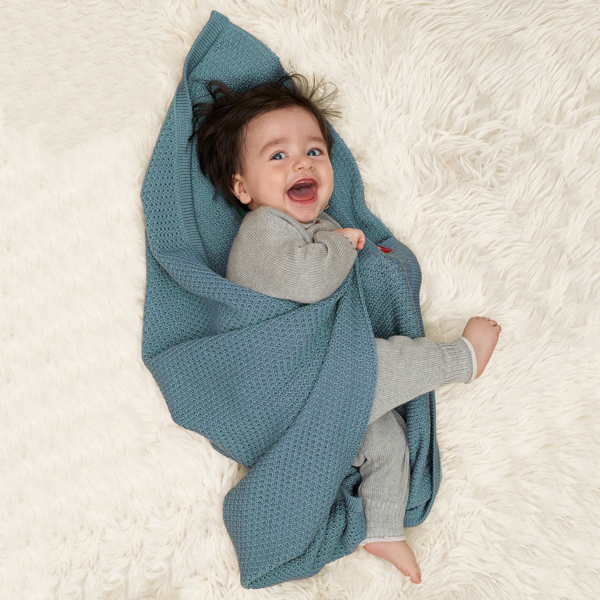 Baby mit dunklen Haaren lacht fröhlich in die Kamera, es hat noch keine Zähne und ist in eine hellblaue gestrickte Babydecke von vincente gefüllt. Die Decke ist aus Merinowolle