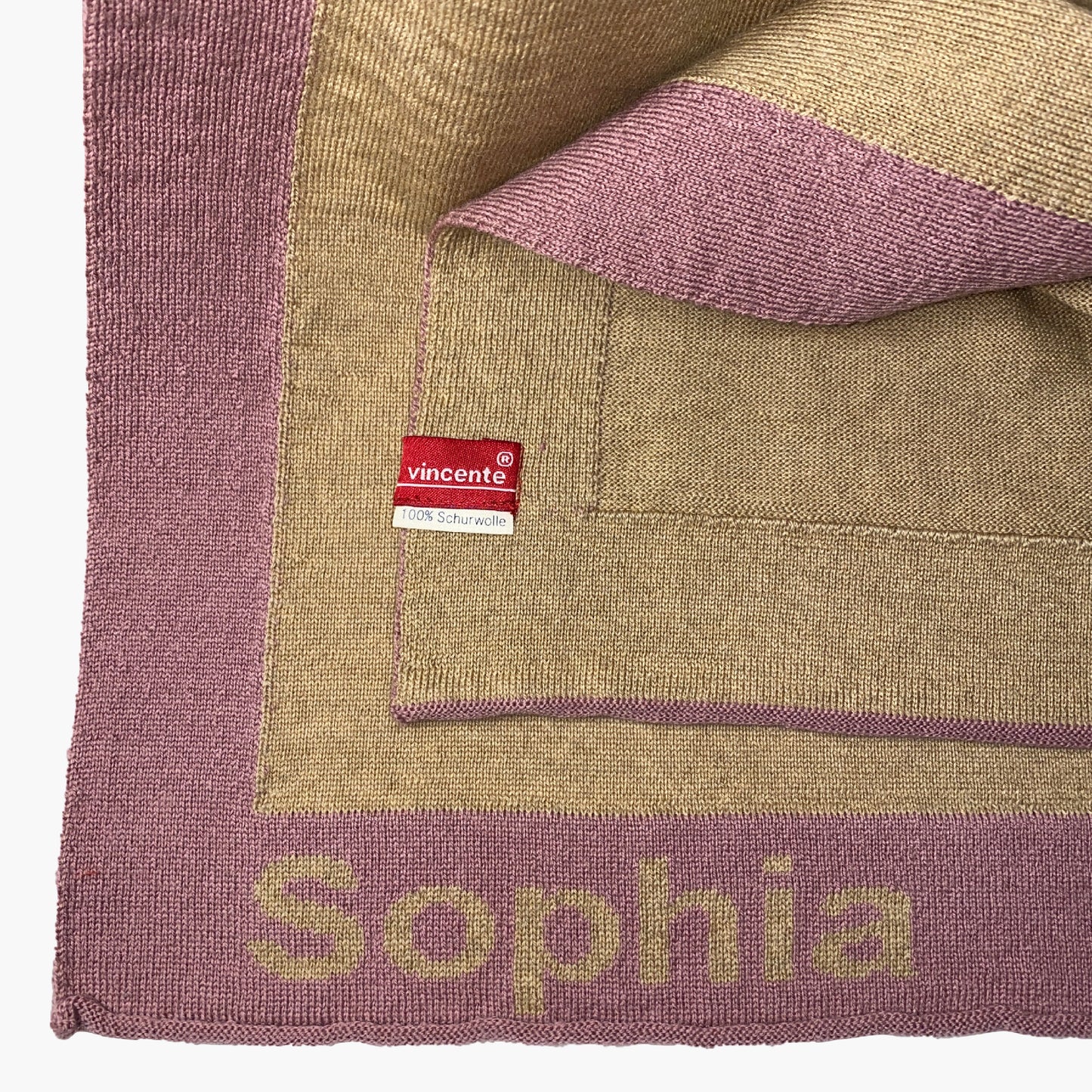 Man sieht eine zusammengefaltete Karamelfarbenen Babydecke. Der Rand ist Rosenholzfarben und mit dem Namen Sophia bestrickt.