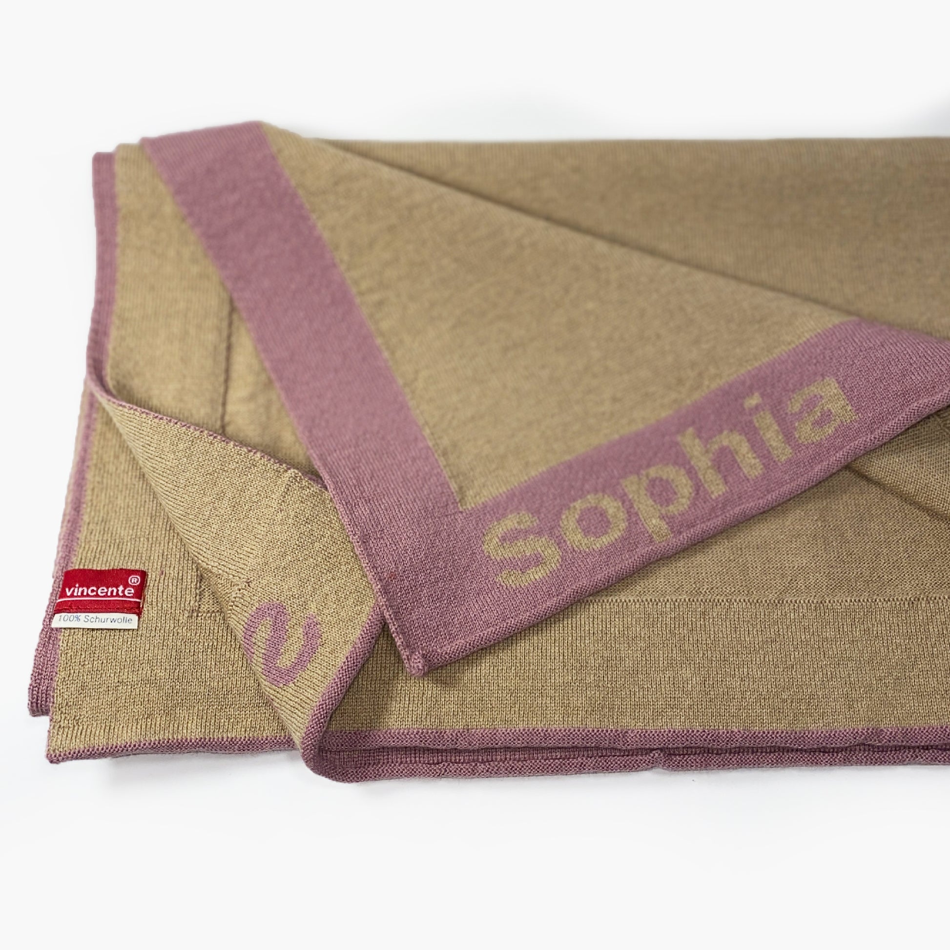 Man sieht eine zusammengefaltete Karamelfarbenen Babydecke. Der Rand ist Rosenholzfarben und mit dem Namen Sophia bestrickt.