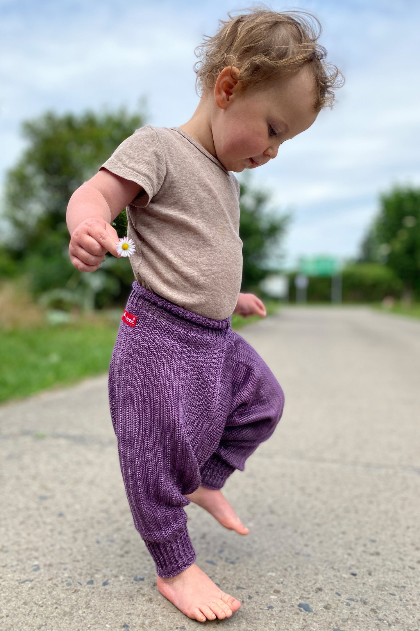 tanzendes Baby mit einem Gänseblümchen in der Hand. Es trägt eine Sarouelhose von vincente aus Tencel/Lyocell gestrickt in Lila