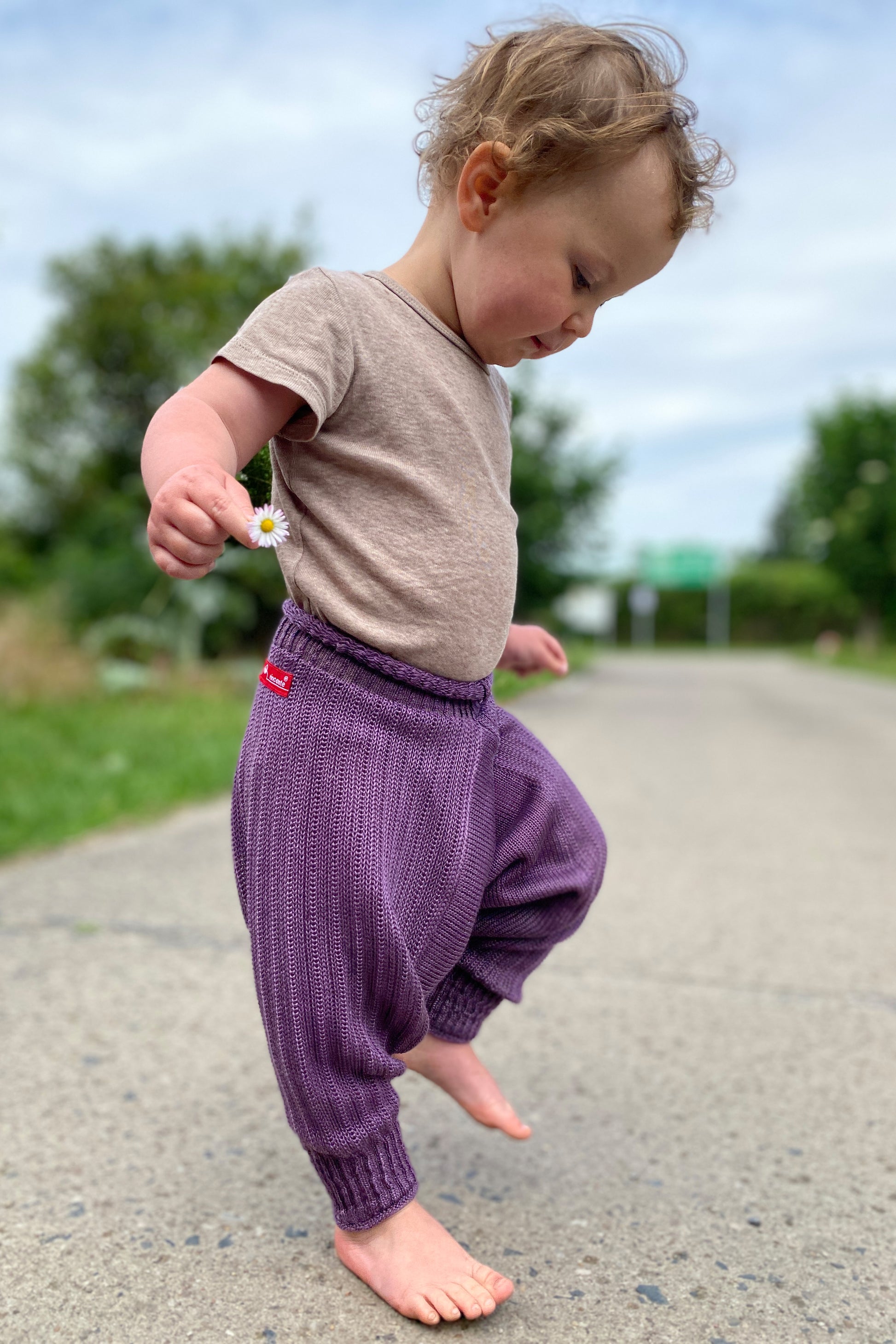 tanzendes Baby mit einem Gänseblümchen in der Hand. Es trägt eine Sarouelhose von vincente aus Tencel/Lyocell gestrickt in Lila