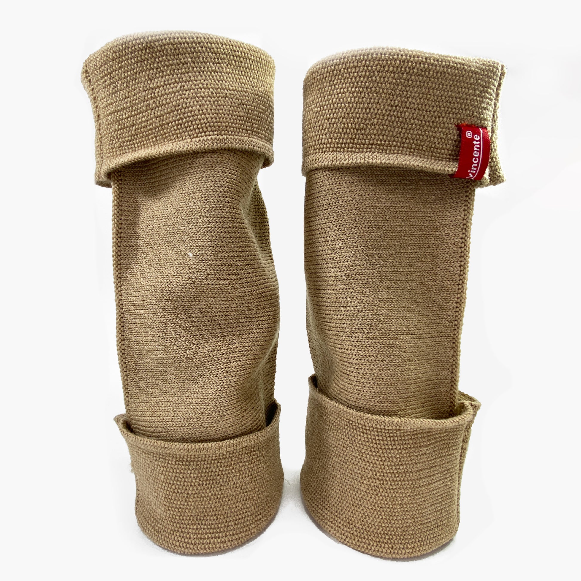 Beige aufrecht stehende Beinstulpen. Man sieht das außenliegende Glatt Rechts Muster. Sie sind umgeschlagen so sieht man das Innere feine Knötchen-Muster. Man kann sie auch Innside-out tragen. Das Rote/Weiße vincente-Logo ist angenäht.