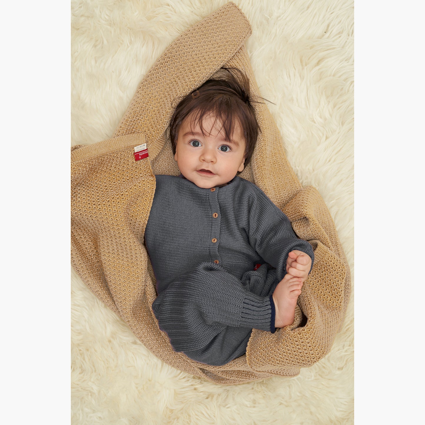 Baby mit dunklen Haaren lacht fröhlich in die Kamera, es hat noch keine Zähne und ist in eine Beigen gestrickte Babydecke von vincente gefüllt. Die Decke ist aus Merinowolle. Ansonsten ist es von oben bis unten mit Strickwaren aus der vincente-Kollektion gekleidet.