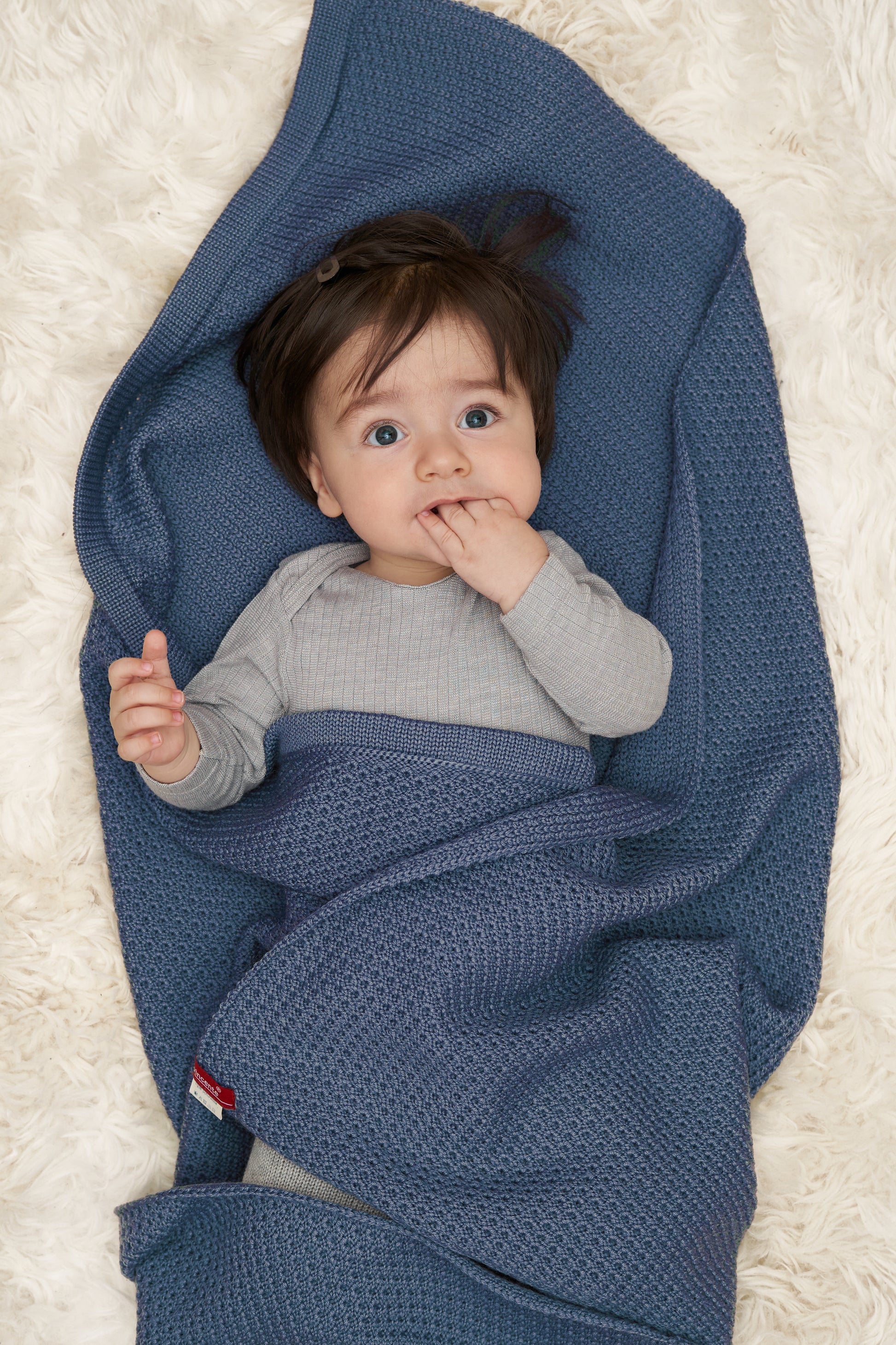 Ein Baby mit dunklen Haaren nuckelt an seinen Fingern, es in eine Blaue, gestrickte Babydecke von vincente gefüllt. Die Decke ist aus Merinowolle. 